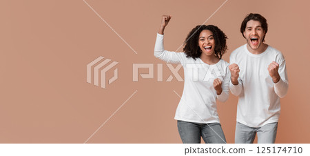 We Did It. Overjoyed multiracial couple celebrating success with raised fists, emotionally reacting to victory, posing together over yellow background 125174710