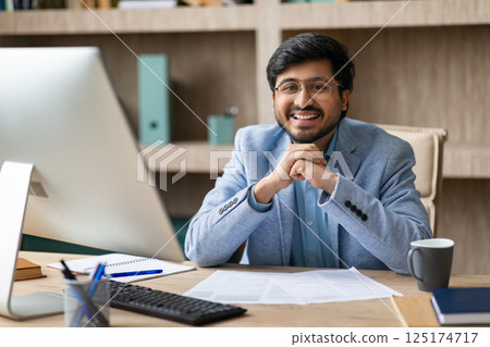 Indian man wearing glasses smiles while sitting at his desk, with a computer and papers in front of him. He has a friendly and confident expression, suggesting he is ready to work or have a meeting. Indian man wearing glasses smiles while sitting at his desk, with a computer and papers in front of him. He has a friendly and confident expression, suggesting he is ready to work or have a meeting. 125174717