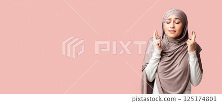 Young muslim girl in hijab crossing fingers, making wish, praying for miracle over pink studio background with free space 125174801
