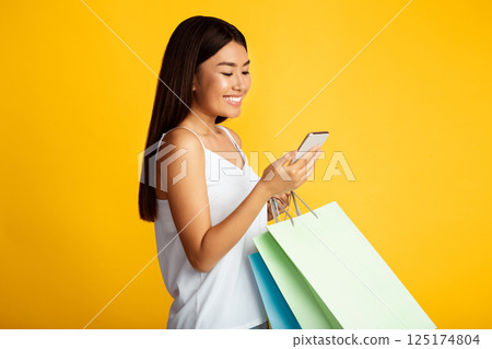 A smiling young Asian woman in a white tank top stands against a yellow background. She is holding a smartphone in her right hand and shopping bags in her left 125174804
