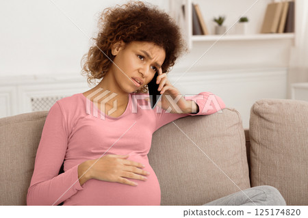 Sad afro pregnant woman talking on phone, having serious conversation with doctor 125174820
