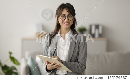 Happy professional middle eastern female psychologist writing in clipboard, looking and smiling at camera, working in modern office. Psychotherapy services, mental health professional concept 125174887