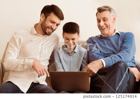 Cheerful Boy Using Laptop Sitting Between Dad And Grandfather On Sofa Indoor. Selective Focus Cheerful Boy Using Laptop Sitting Between Dad And Grandfather On Sofa Indoor. Selective Focus 125174914
