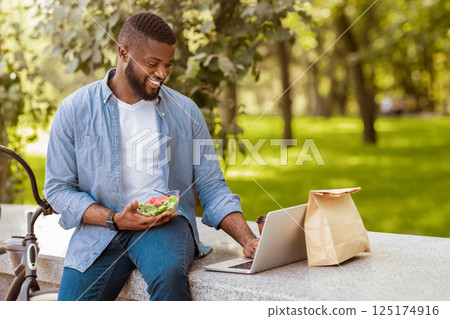 Happy afro businessman using laptop and having healthy lunch outdoors with bicycle parked nearby Happy afro businessman using laptop and having healthy lunch outdoors with bicycle parked nearby 125174916