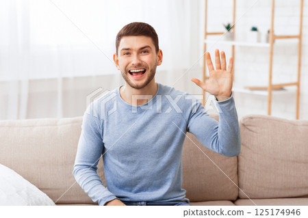 Hello. Cheerful Guy Waving Hand Smiling Looking At Camera Sitting On Couch Indoor. Hello. Cheerful Guy Waving Hand Smiling Looking At Camera Sitting On Couch Indoor. 125174946