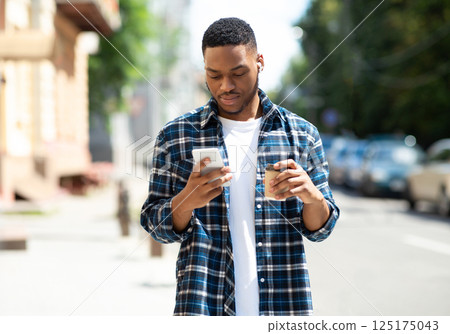 Good-looking young black hipster messaging online on his cell phone, walking down the street with coffee cup 125175043