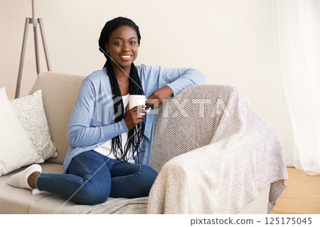 Portrait of attractive african american woman sitting on sofa with cup of coffee and smiling at camera, free space 125175045