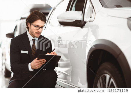 Writing Report. Serious handsome auto insurance agent man in glasses filling insurance form sitting near used car 125175129