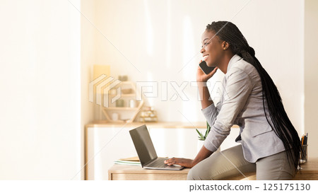Business Communication. Young Black Businesswoman Talking On Cellphone And Using Laptop, Leaning On Her Desk In Office, Side View Business Communication. Young Black Businesswoman Talking On Cellphone And Using Laptop, Leaning On Her Desk In Office, Side View 125175130