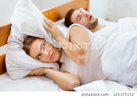 Snoring Concept. Irritated Woman Covering Ears With Pillow Lying Near Sleeping Husband Unable To Sleep In Bed Indoor. Selective Focus Snoring Concept. Irritated Woman Covering Ears With Pillow Lying Near Sleeping Husband Unable To Sleep In Bed Indoor. Selective Focus 125175132