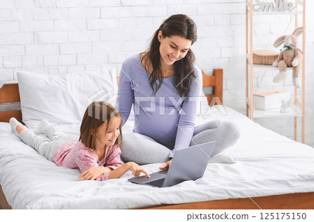 Little girl and pregnant woman using laptop, sitting on bed and having fun together at home on weekend, copy space 125175150