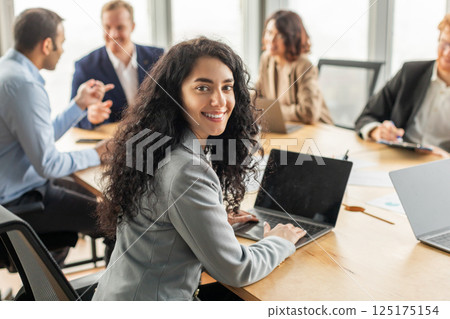 A young businesswoman sits at a conference table with colleagues, working on a laptop. She smiles and looks over her shoulder at the camera while typing on her computer with blank screen A young businesswoman sits at a conference table with colleagues, working on a laptop. She smiles and looks over her shoulder at the camera while typing on her computer with blank screen 125175154
