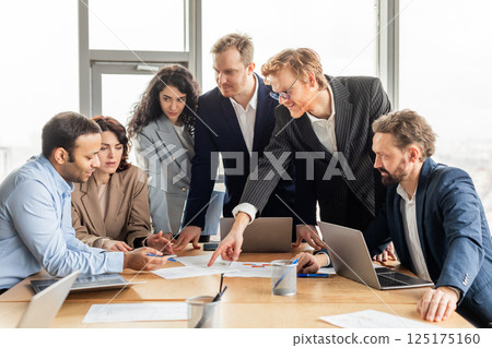 A group of business professionals are gathered around a table in an office setting, discussing a project. The team is reviewing documents and using laptops as they brainstorm ideas and strategize 125175160
