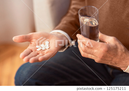 Unrecognizable Senior Gentleman Holding Pills And Water Sitting On Sofa At Home. Cropped, Selective Focus Unrecognizable Senior Gentleman Holding Pills And Water Sitting On Sofa At Home. Cropped, Selective Focus 125175211