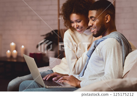 Excited afro couple surfing net on laptop enjoying cozy winter atmosphere at home, free space Excited afro couple surfing net on laptop enjoying cozy winter atmosphere at home, free space 125175212