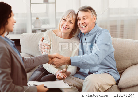 Sealing A Deal. Happy elderly couple sitting on couch and shaking hands with young female financial advisor 125175244