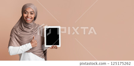 Great App. Positive black islamic girl in hijab holding digital tablet with black screen and showing thumb up gesture over yellow background, mockup 125175262