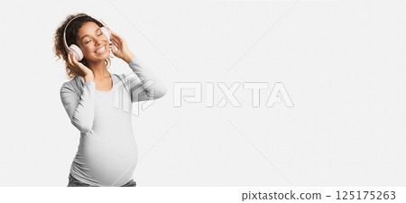 Afro pregnant woman enjoying music in headphones with closed eyes, white studio background 125175263