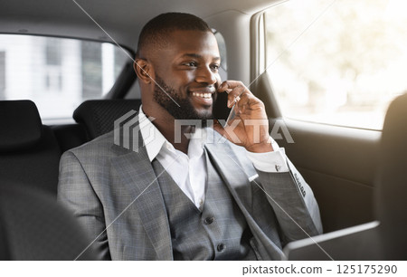 Happy black businessman with laptop talking on mobile phone with clients, looking through window, going to business meeting by car, african manager sitting on back seat, having conversation on phone 125175290