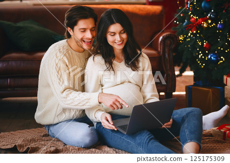 Pregnant cheerful lady using laptop with her husband on Christmas Eve, sitting by xmas tree at home, shopping online, copy space Pregnant cheerful lady using laptop with her husband on Christmas Eve, sitting by xmas tree at home, shopping online, copy space 125175309