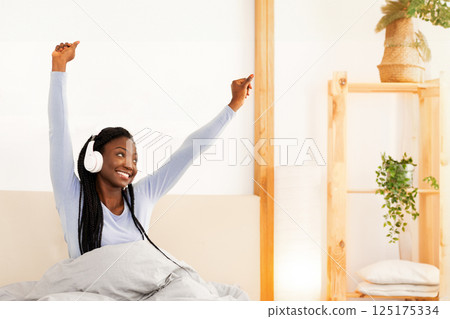 Good Morning. Happy Afro Lady In Headphones Stretching Arms Sitting In Cozy Bed At Home. Free Space 125175334