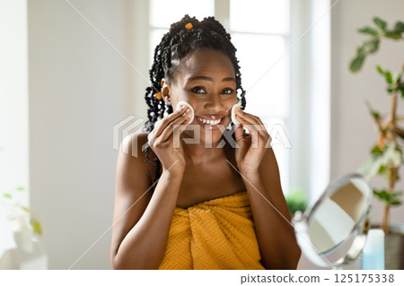 African American woman with braided black hair is wrapped in a yellow towel and smiling at the camera while using a cotton round on her cheek. African American woman with braided black hair is wrapped in a yellow towel and smiling at the camera while using a cotton round on her cheek. 125175338