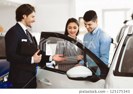 Auto Business Concept. Handsome Assistant Presenting New Luxury Auto With Open Door To Young Couple Auto Business Concept. Handsome Assistant Presenting New Luxury Auto With Open Door To Young Couple 125175343