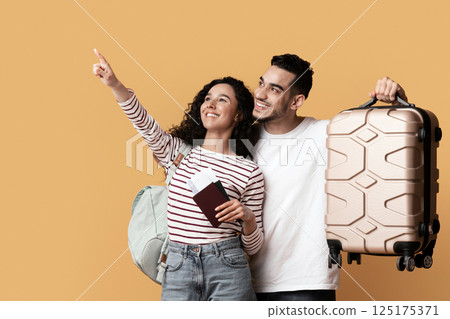 Travel Advertisement. Joyful Arab Couple With Suitcase And Passports Pointing Away At Copy Space On Yellow Background, Happy Middle Eastern Travellers Demonstrating Free Place For Ad, Panorama 125175371