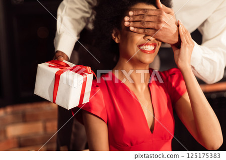 Romantic Surprise. Unrecognizable Afro Guy Giving Valentine Gift To Girlfriend Covering Her Eyes Celebrating In Restaurant. Cropped Romantic Surprise. Unrecognizable Afro Guy Giving Valentine Gift To Girlfriend Covering Her Eyes Celebrating In Restaurant. Cropped 125175383