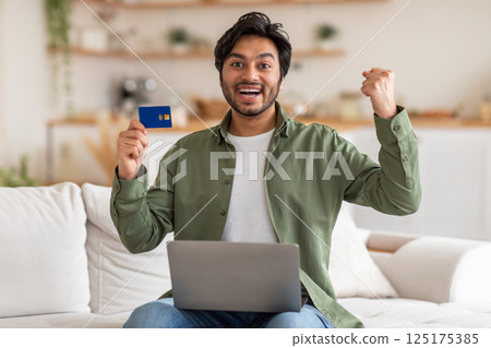 A young Arab man sits on a white couch, holding a credit card in one hand and raising his other fist in the air in celebration. He is smiling excitedly, with a laptop resting on his lap. 125175385