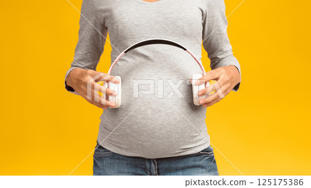 Music and pregnancy. Expectant woman holding headphones near belly, pink studio background, crop 125175386