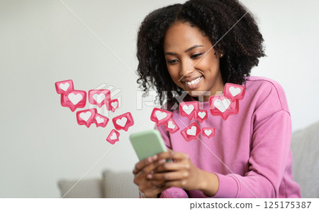 A young black woman with curly hair sits on a couch and smiles as she looks at a smartphone, surrounded by small, pink icons with white hearts inside. 125175387