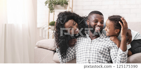 Happy Father's Day Concept. African american man enjoying spending time with wife and daughter at home, enbracing and laughing together 125175390