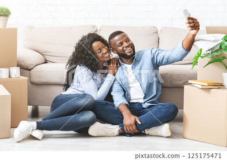 New life in new house. Young happy african american couple embracing at new apartment, taking selfie on smartphone, copy space New life in new house. Young happy african american couple embracing at new apartment, taking selfie on smartphone, copy space 125175471