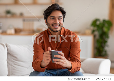 Arab man in an orange shirt sits on a white couch in his living room, smiling and holding a grey mug in his hands. He looks off to the side, likely deep in thought or enjoying a moment of peace 125175475