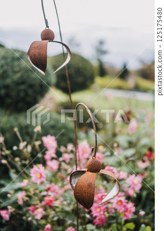 Rustic metal angel sculptures hanging in a flower garden with pink blossoms. Garden art, symbolic decor, spiritual aesthetics, handmade craftsmanship 125175480
