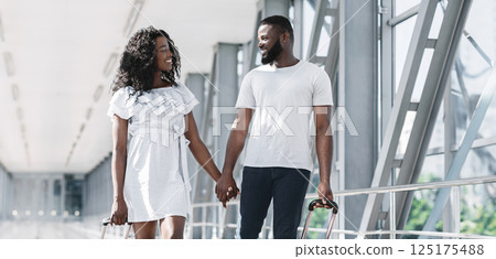 A young black couple walks hand-in-hand through a modern airport terminal, each pulling a rolling suitcase behind them. They are smiling and seem to be enjoying their time together, copy space A young black couple walks hand-in-hand through a modern airport terminal, each pulling a rolling suitcase behind them. They are smiling and seem to be enjoying their time together, copy space 125175488