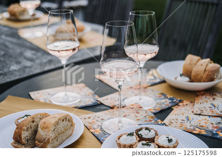 Glasses of rose wine and savory tarts arranged on an outdoor table set with decorative napkins and fresh bread. Al fresco dining, artisanal food, celebration gathering, slow living 125175598