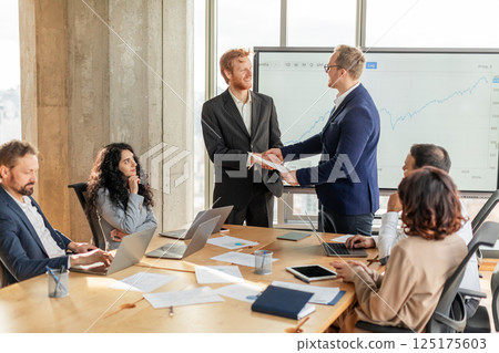 A group of business professionals are gathered around a large conference table. Two men in suits are shaking hands while standing, signaling a successful agreement or partnership 125175603