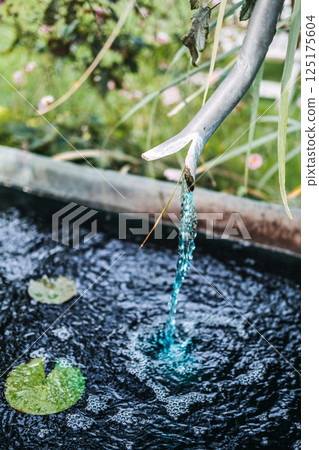 Water flows from a sculpted metal pipe into a tranquil garden pond with lily pads. Water conservation, eco-friendly garden design, artistic landscaping, sustainability 125175604