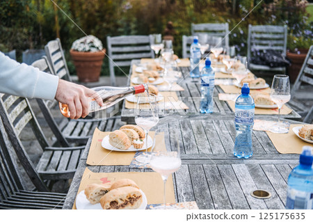 A hand pours rose wine into a glass on a rustic outdoor table set with bread, tarts, and bottled water. Minimal menu, grounded luxury, artisanal wine, water and wine culture A hand pours rose wine into a glass on a rustic outdoor table set with bread, tarts, and bottled water. Minimal menu, grounded luxury, artisanal wine, water and wine culture 125175635
