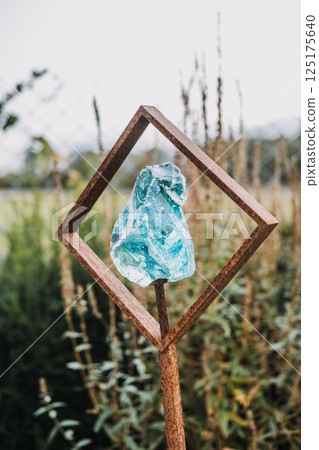Aqua-colored glass sculpture held within a rusty iron frame displayed in a garden setting. This image expresses modern outdoor decor, raw materials, earth energy, and art in nature Aqua-colored glass sculpture held within a rusty iron frame displayed in a garden setting. This image expresses modern outdoor decor, raw materials, earth energy, and art in nature 125175640