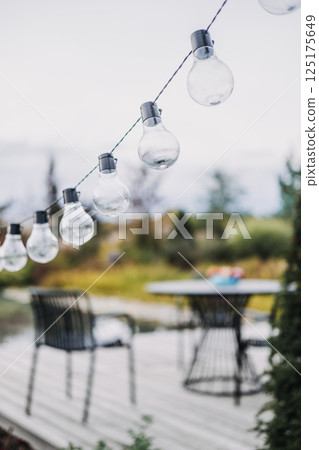 String of clear outdoor bulbs hanging above a garden patio with modern furniture in soft focus. Ambient garden lighting, outdoor entertaining, minimalist backyard design, cozy atmosphere 125175649