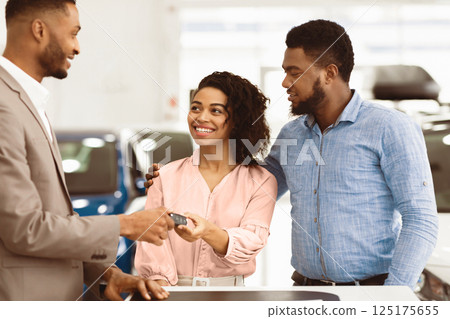 Car Dealer Giving Key To Cheerful Afro Couple Buyers Standing In Auto Dealership Center. Cars Sales. Selective Focus 125175655