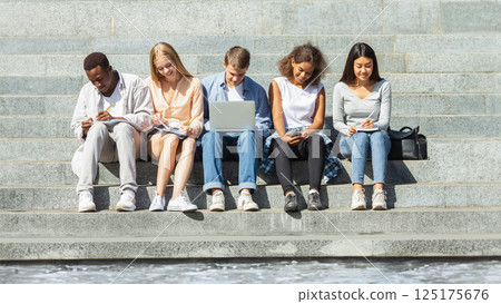 Multiethnic group of students preparing for exams, studying in park, using laptop, phone, tablet 125175676
