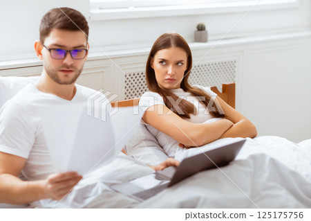 Modern Relationship Problems. Young Husband Working On Laptop Lying With Angry Discontented Wife In Bed At Home. Selective Focus 125175756