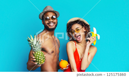 Black man and woman are smiling and posing for the camera against a blue background. The man is wearing sunglasses and a hat and holding a pineapple. The woman is holding a banana and an orange. 125175860