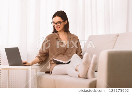 Knowledge Concept. Relaxed girl in glasses sitting on couch, reading book, holding notebook and using laptop, copyspace Knowledge Concept. Relaxed girl in glasses sitting on couch, reading book, holding notebook and using laptop, copyspace 125175902