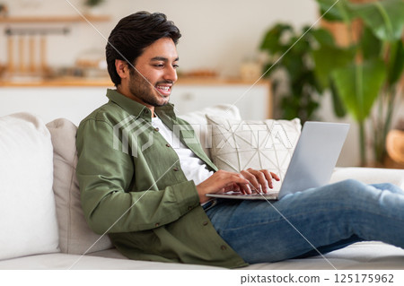 Arab man is sitting on a couch inside a home. He is smiling and using a laptop, appearing relaxed and comfortable. The man is wearing casual clothing, including a green shirt and blue jeans. 125175962