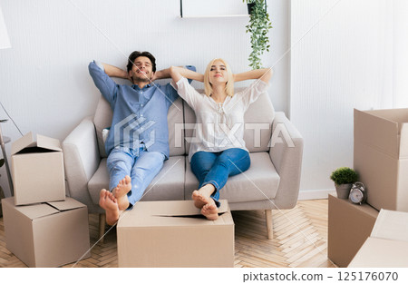 A relaxed couple sits on a couch in their new home, surrounded by cardboard boxes. 125176070